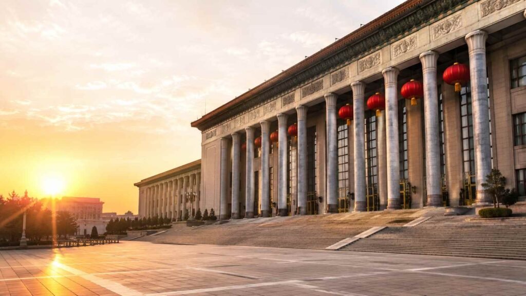 Wide-angle exterior view of the Great Hall of the People in Beijing at dawn with red lanterns, stone steps, and soft golden morning light in a clean editorial news-style scene without people